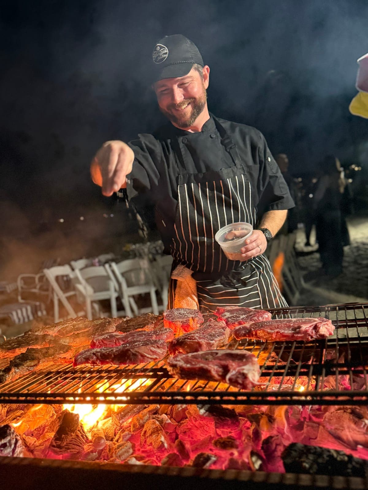 Chef Matt Brophy seasoning steaks over a wood fire grill at a San Diego beach party