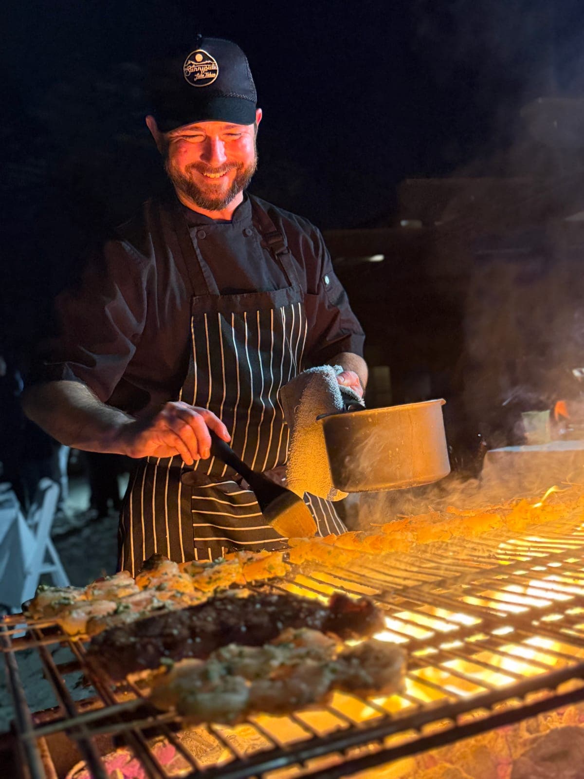 Chef Matt Brophy grilling shrimp and steak over open flame at a private beach event in San Diego